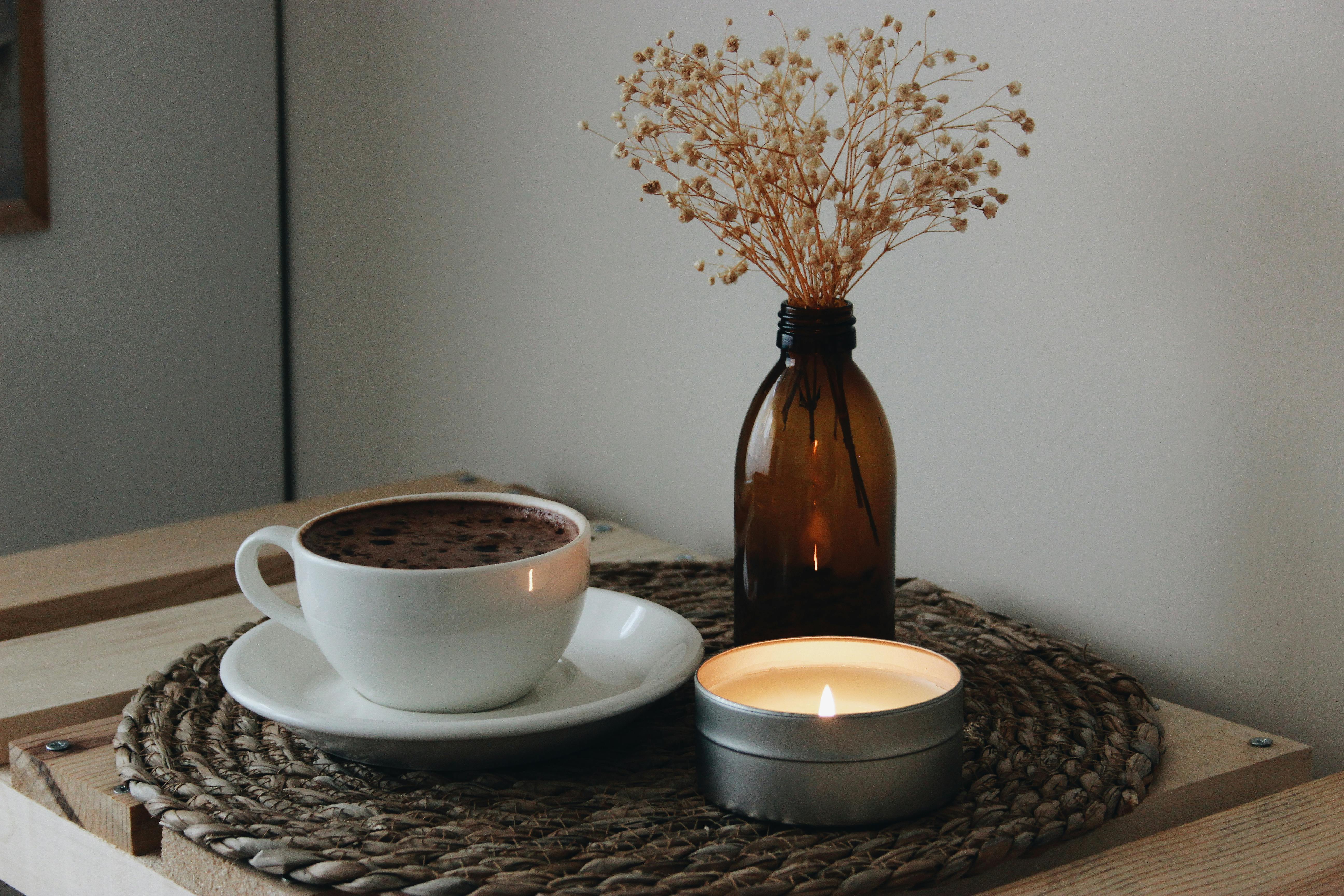 Candle burning, hot tea in a mug, and vase sitting on table.